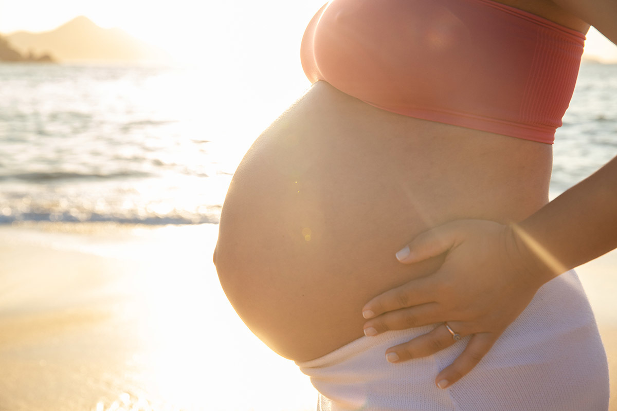 Detalhe da barriga da gestante com o mar ao fundo e o sol radiante na Praia Vermelha.