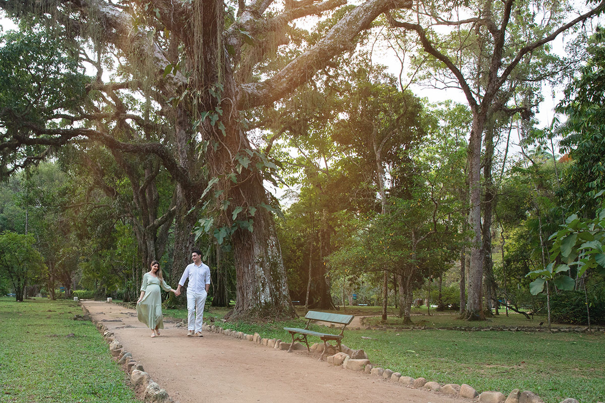 Casal sorridente em ensaio de gestação no Jardim Botânico, caminhando juntos de mãos dadas.
