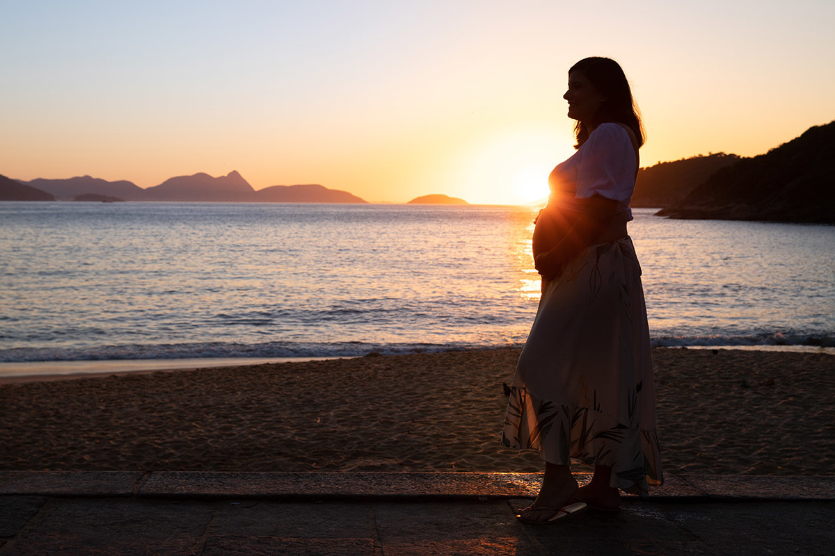 Foto de gestante na Praia Vermelha, luz do nascer do sol iluminando a barriga.