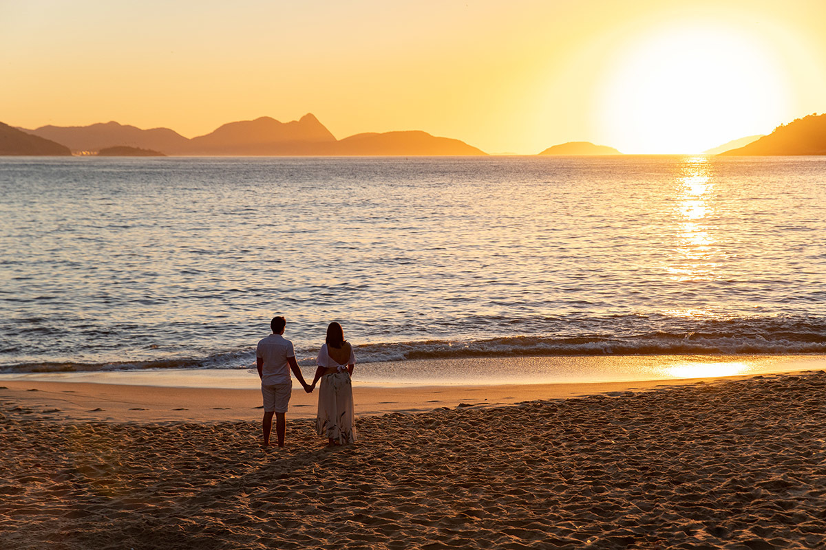 Casal em ensaio de gestante na Praia Vermelha, na areia com cenário do mar ao fundo.
