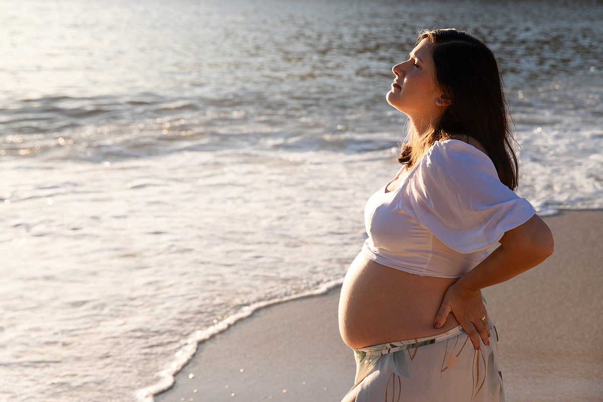 Foto de gestante na Praia Vermelha, luz do nascer do sol iluminando a barriga.