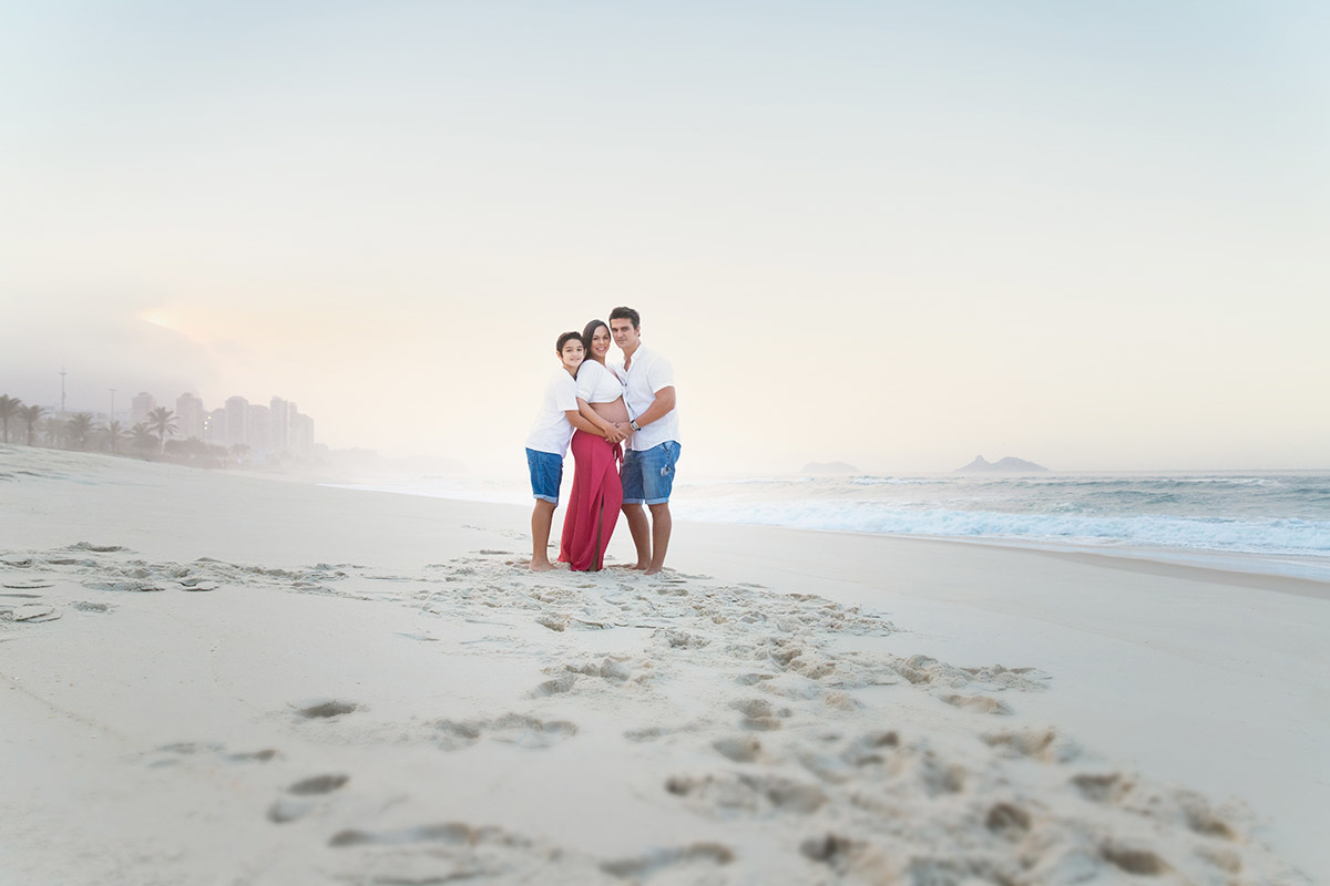 Ensaio de gestante na praia: Silvia, Sanderson e Matheus abraçados, no Rio de Janeiro.