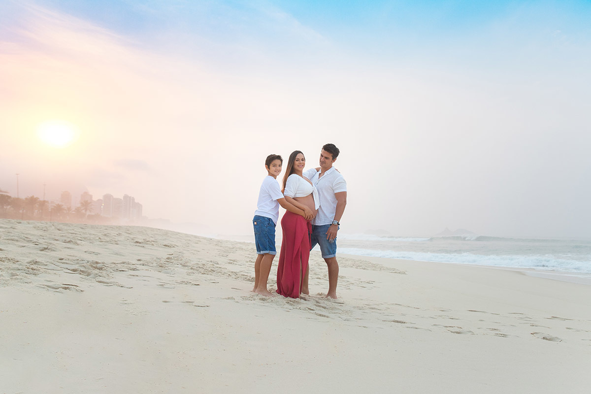 Ensaio de gestante na praia: Silvia, Sanderson e Matheus abraçados, no Rio de Janeiro.