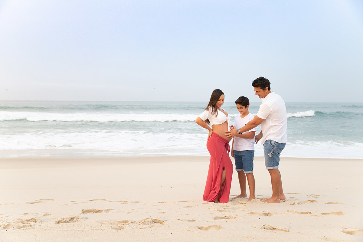 Ensaio de gestante na praia: Silvia, Sanderson e Matheus abraçados, no Rio de Janeiro.