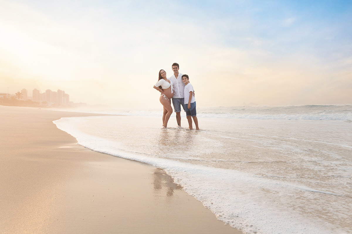 Ensaio de gestante na praia: Silvia, Sanderson e Matheus abraçados, no Rio de Janeiro.