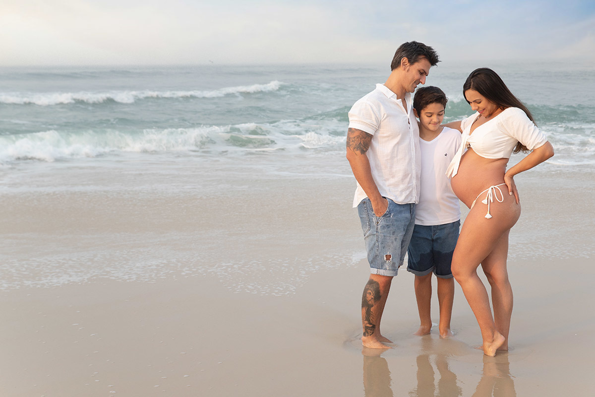Ensaio de gestante na praia: Silvia, Sanderson e Matheus abraçados, no Rio de Janeiro.