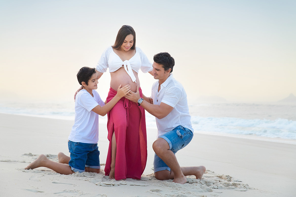 Ensaio de gestante na praia: Silvia, Sanderson e Matheus abraçados, no Rio de Janeiro.