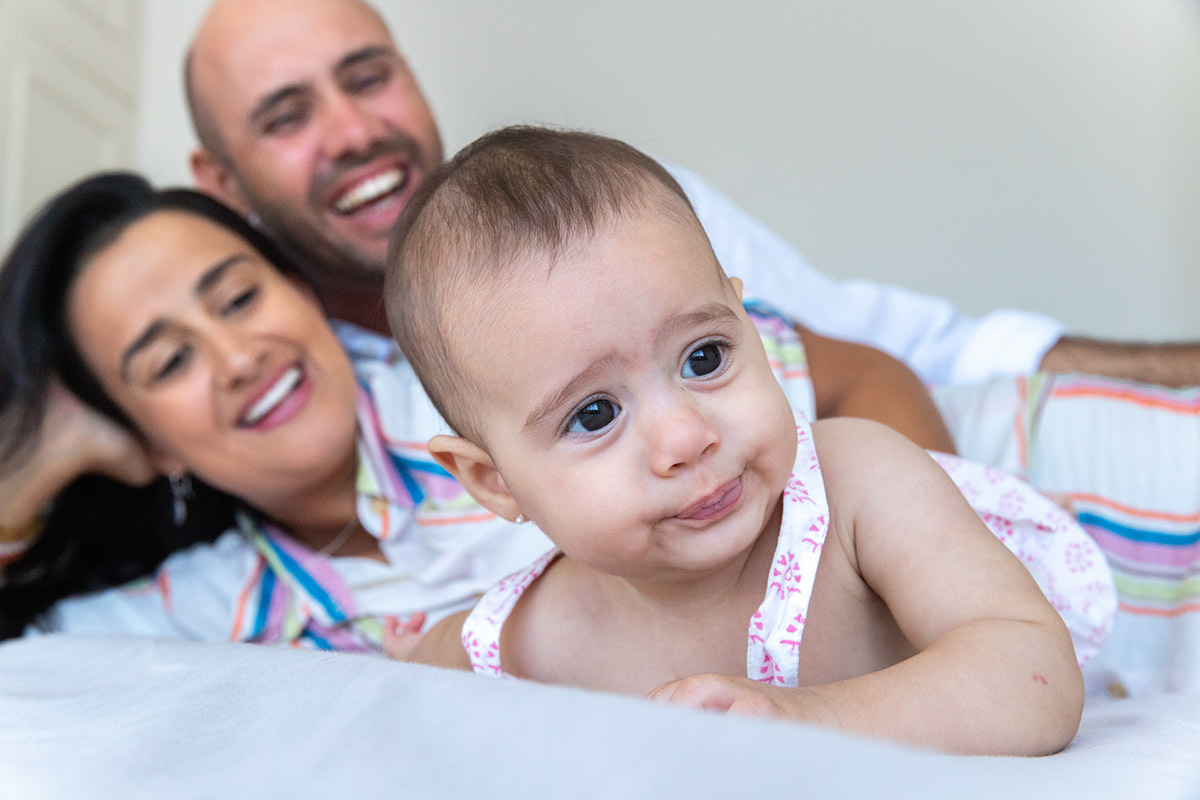 Ensaio de família: Marina deitada na cama com os pais sorrindo, no Rio de Janeiro.