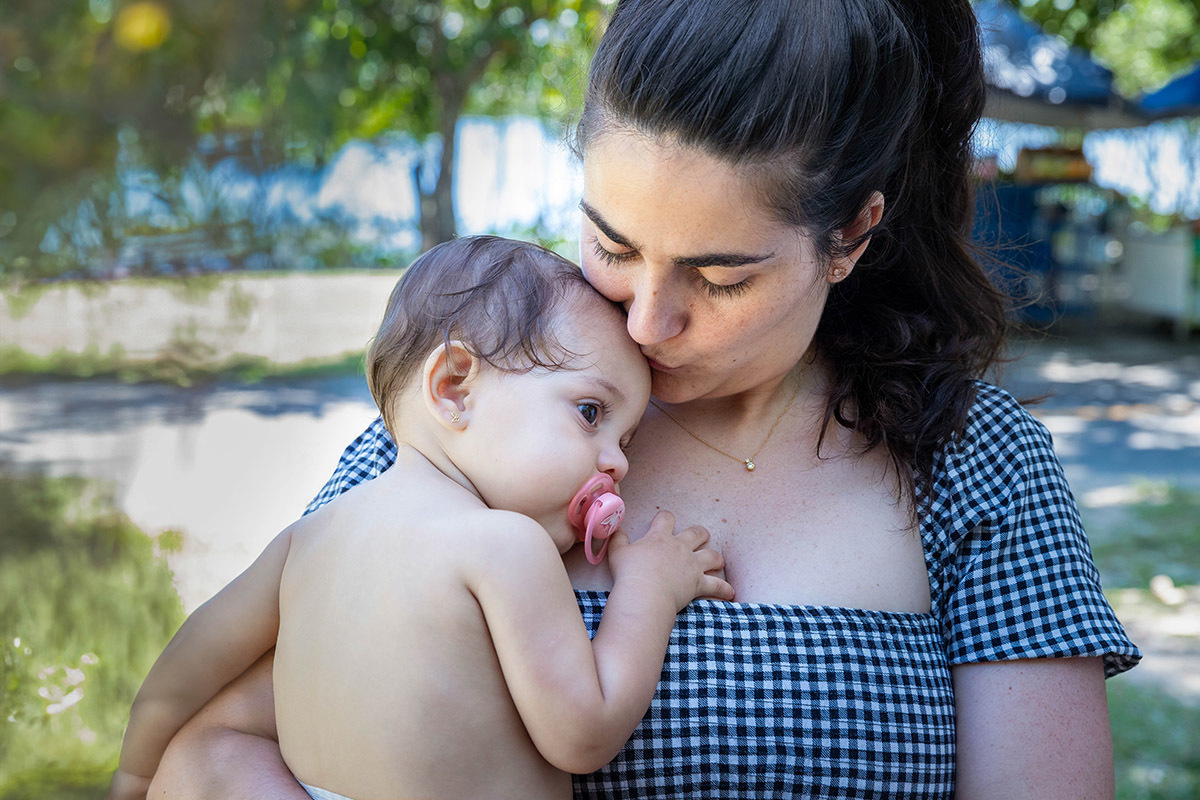 Alice no colo da mãe que está dando um beijo em sua testa durante a festa na Lagoa