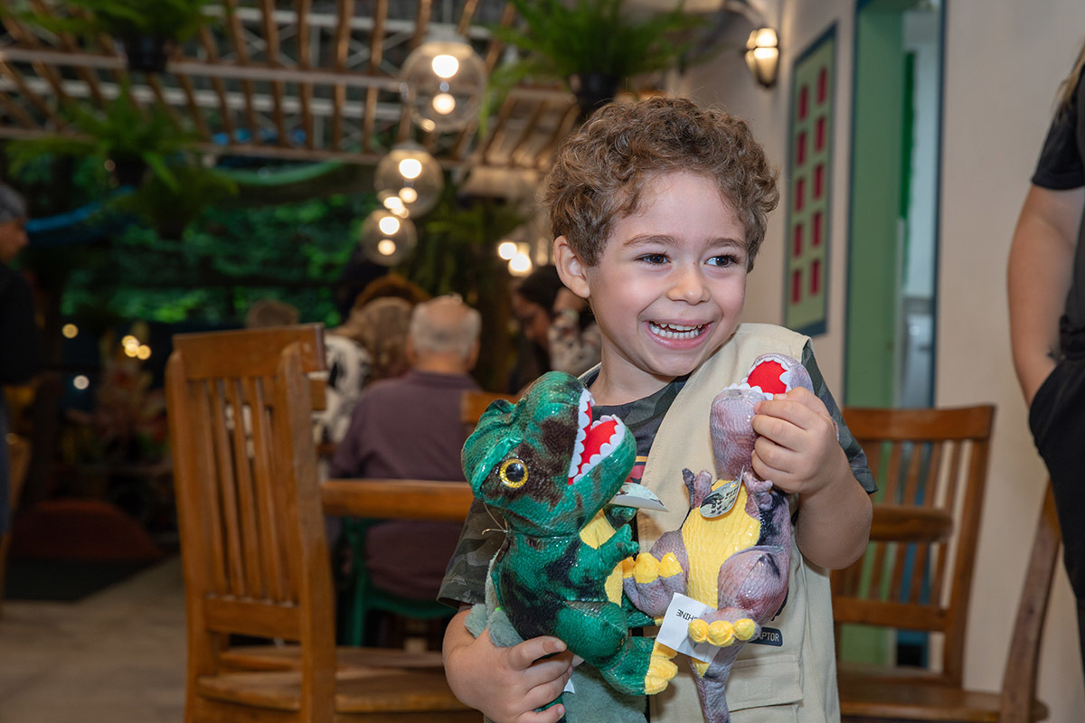 aniversariante sorrindo com dinossauros de pelúcia na festa infantil no espaço aldeia