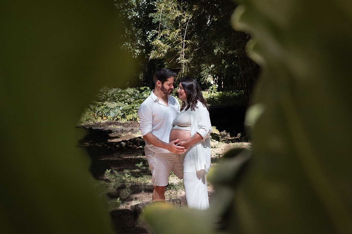 casal de gestante abraçados emoldurados por uma planta durante ensiao no Jardim Botânico