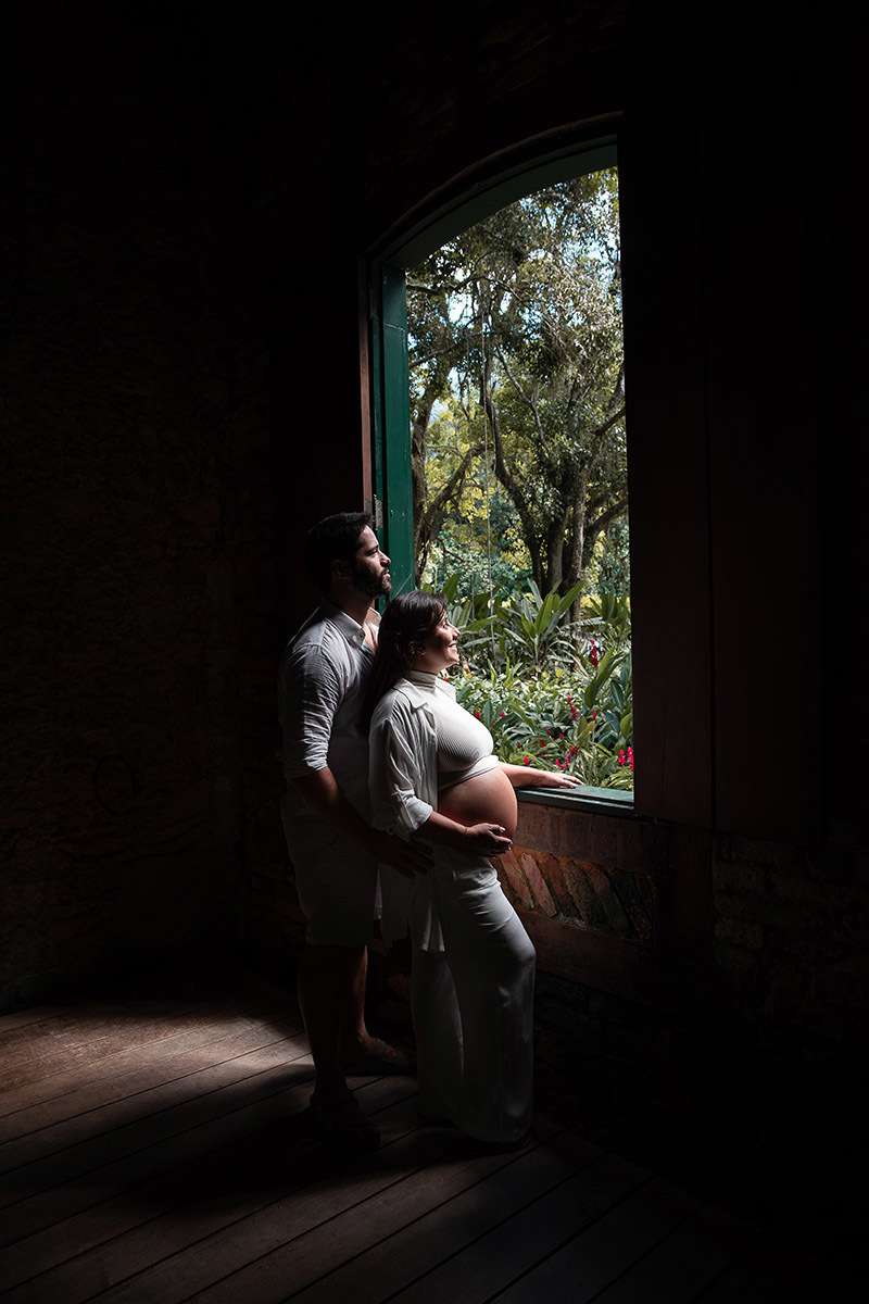 Casal de gestante abraçados na janela olhando para as plantas do jardim Botânico