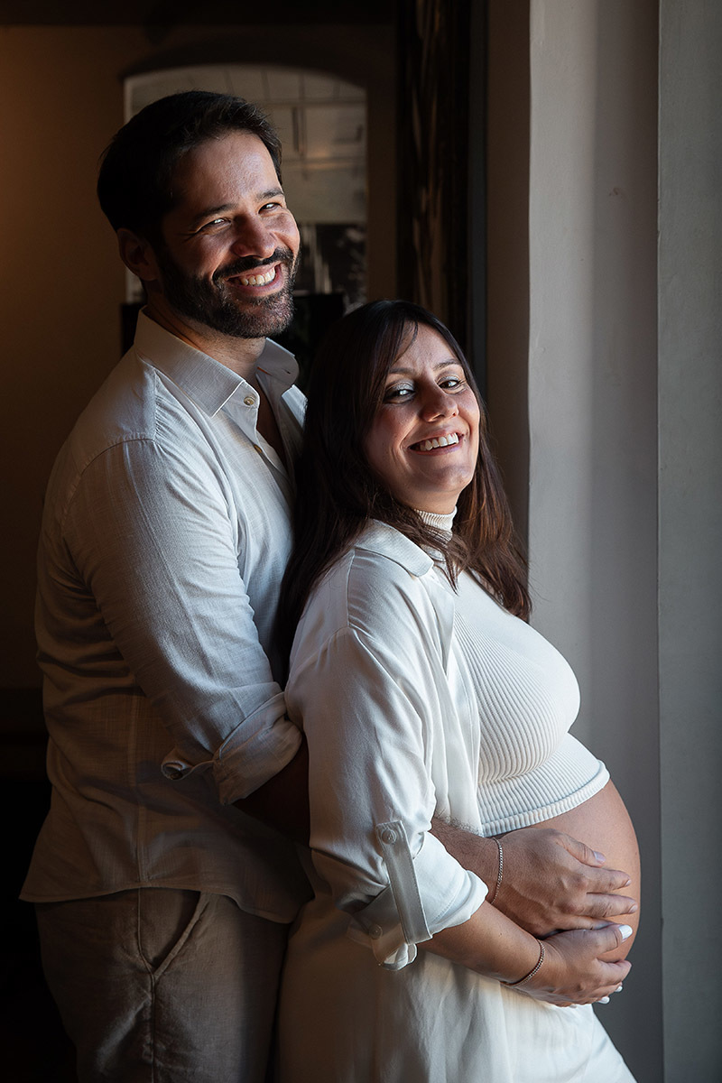 casal de gestante sorrindo e se abraçando com a luz da janela 