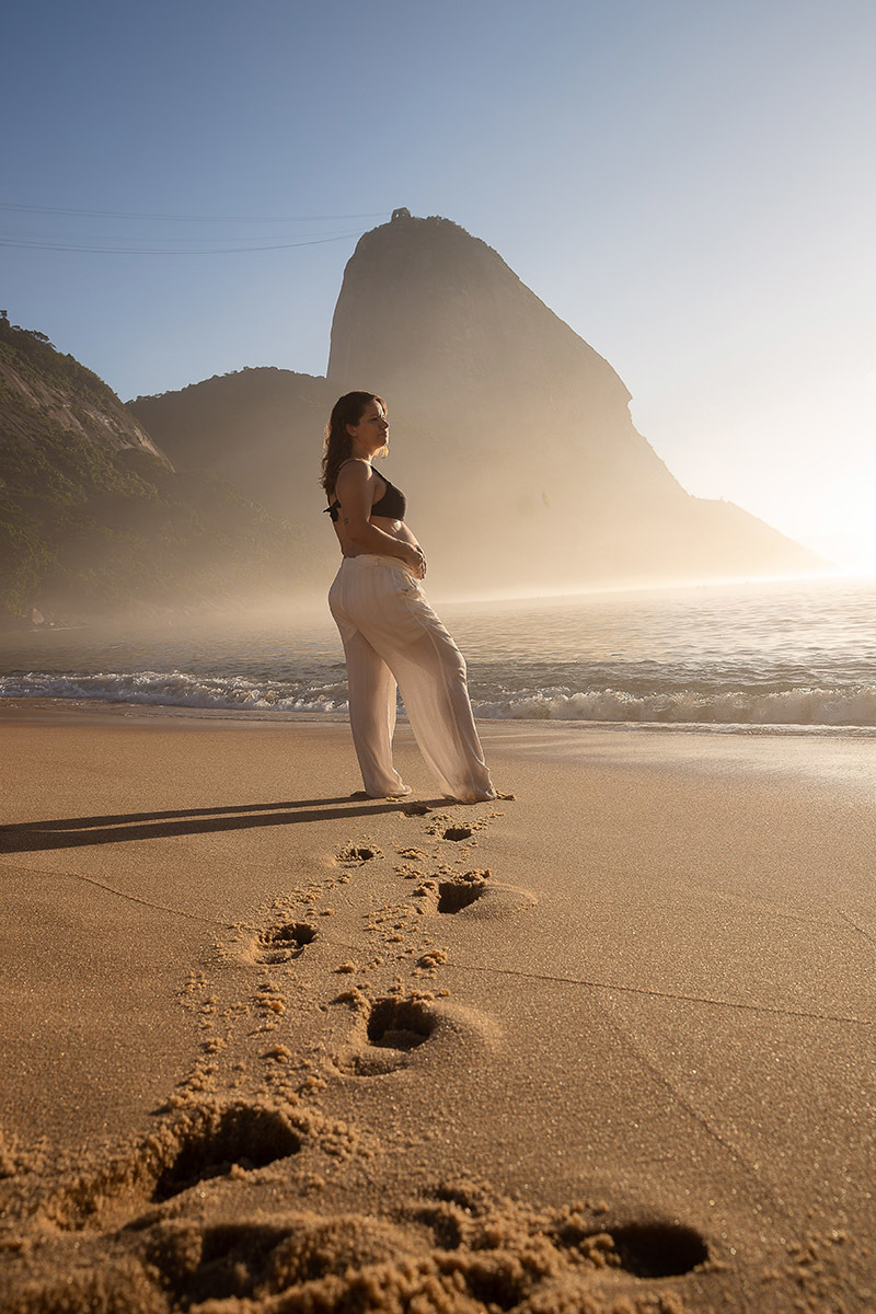 grávida contempla o mar na Praia Vermelha durante o nascer do sol, com o Morro da Urca ao fundo.