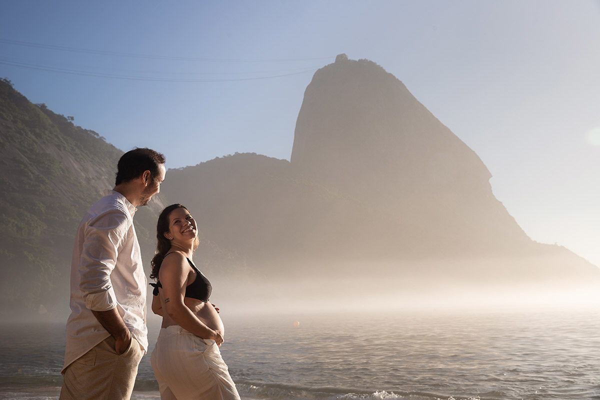 casal se olha de maneira romântica durante ensaio na Praia Vermelha com o mar e morra da Urca ao fundo