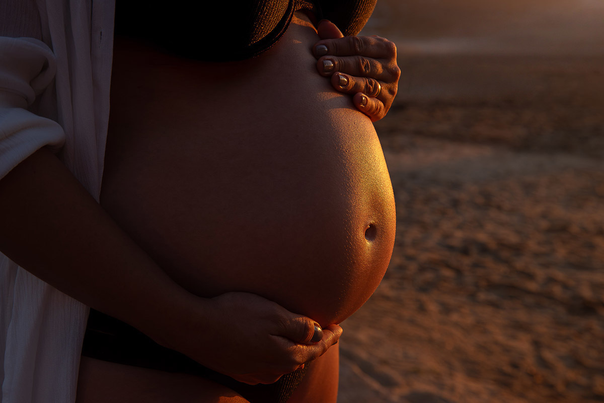 detalhe da barriga da gestante com luz dourada do nascer do sol durante ensaio na Praia Vermelha