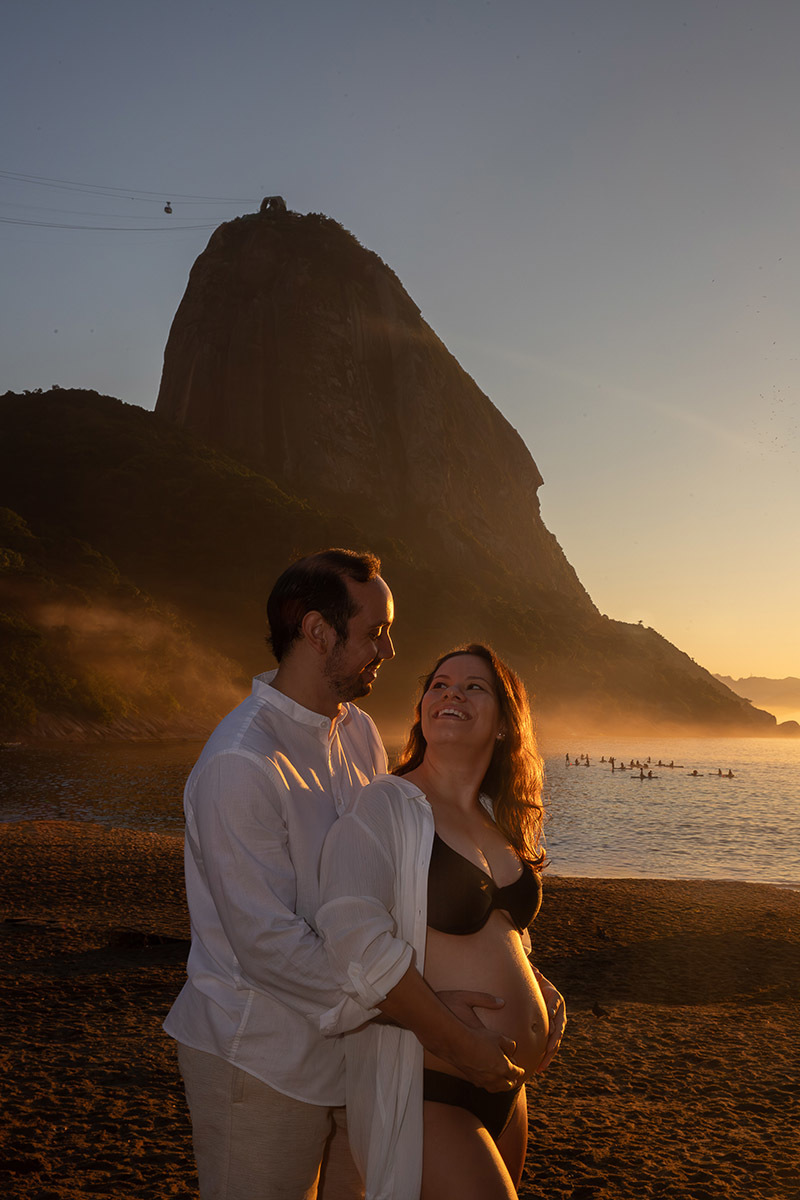 Casal de gestante se olhando e rindo com névoa e luz dourada durante ensaio na Praia Vermelha