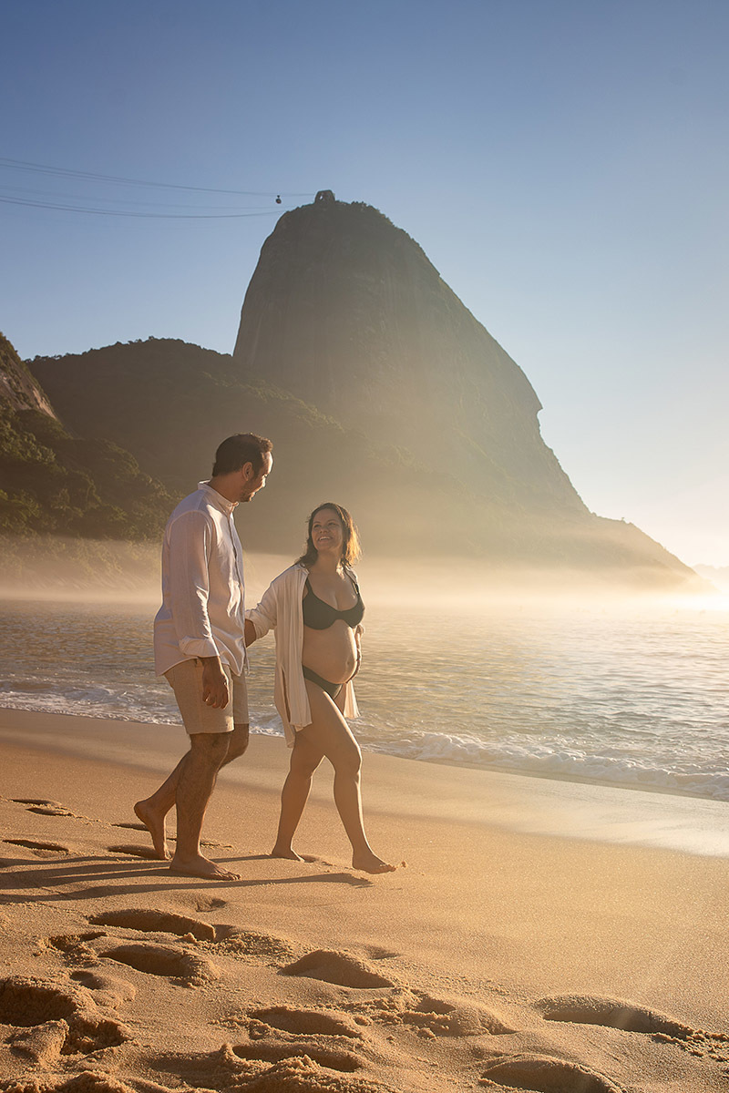 Casal  caminha juntos pela areia da Praia Vermelha durante o ensaio de gestante, com o Morro da Urca ao fundo.
