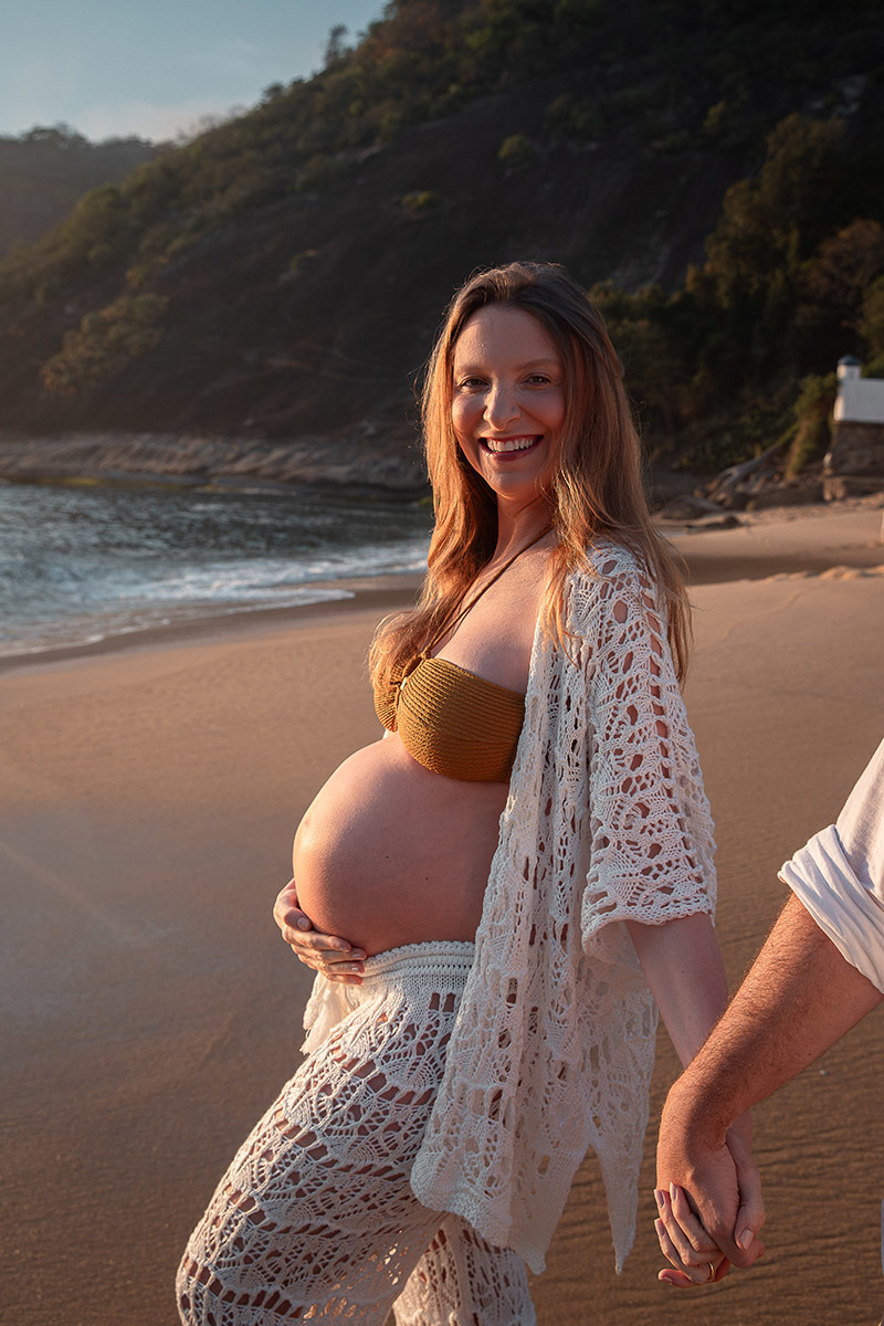 Gestante sorrindo com a mão na barriga e mão dada com o marido durante ensaio na Praia Vermelha