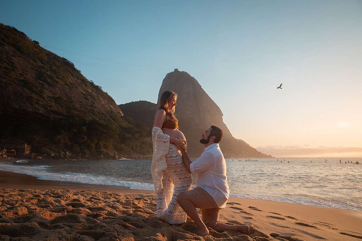Marido ajoelhado olhando para a gestante com olhar de admiração na Praia Vermelha
