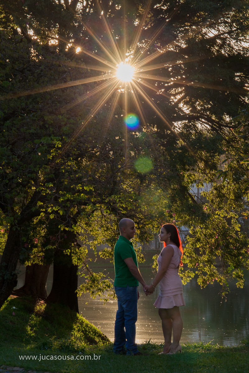 Casal em corpo inteiro com árvores ao fundo e o vazar dos raios solares por entre as folhas formando uma bela estrela e realçando o cabelo de cor vermelha da noiva
