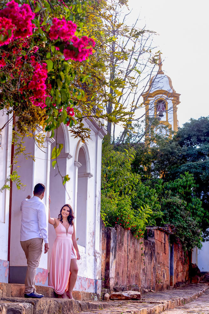 foto tirada em Tiradentes MInas Gerais onde a noiva esta de frente e o noivo de costas encostados na porta de um casarão histórico com a igreja matriz ao fundo