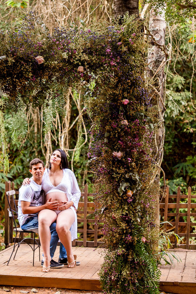 ensaio embaixo de várias arvores externo de gestante no Bouganville Farm gestante sentada no colo do marido