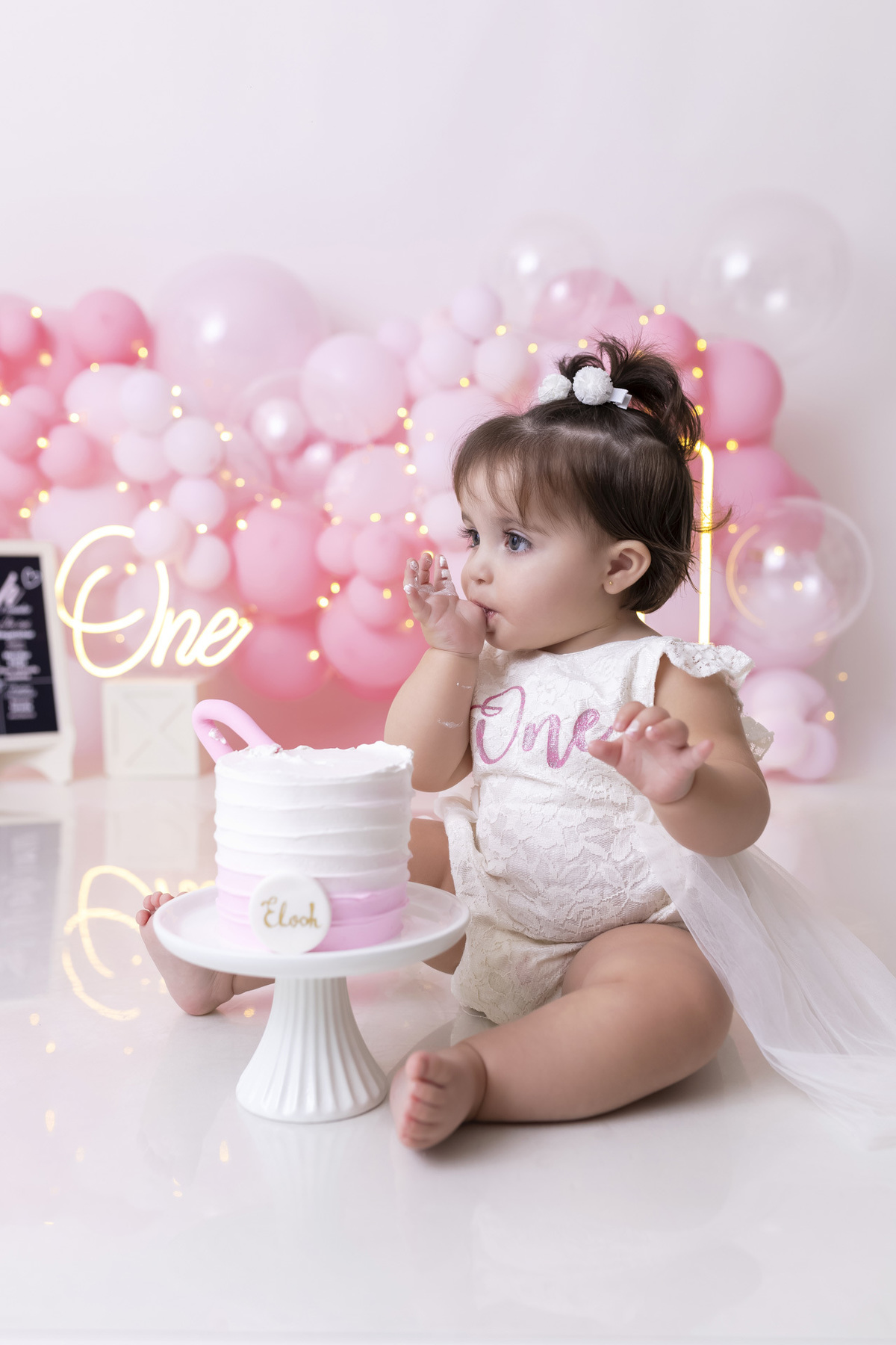 Bebê em ensaio Smash the Cake tema princesa rosa em estúdio fotográfico em Caraguatatuba, cenário delicado, vestido rosa, bolo personalizado e criança sorrindo enquanto brinca com o bolo