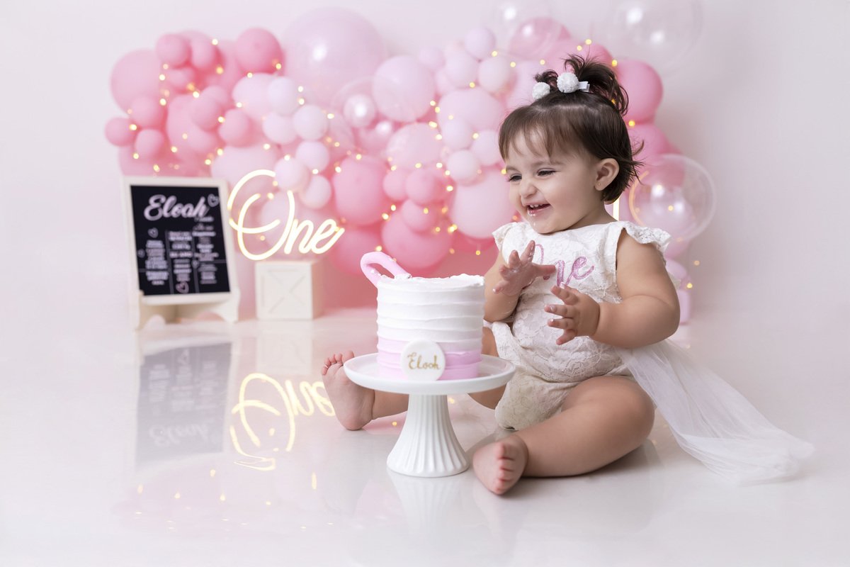 Bebê em ensaio Smash the Cake tema princesa rosa em estúdio fotográfico em Caraguatatuba, cenário delicado, vestido rosa, bolo personalizado e criança sorrindo enquanto brinca com o bolo