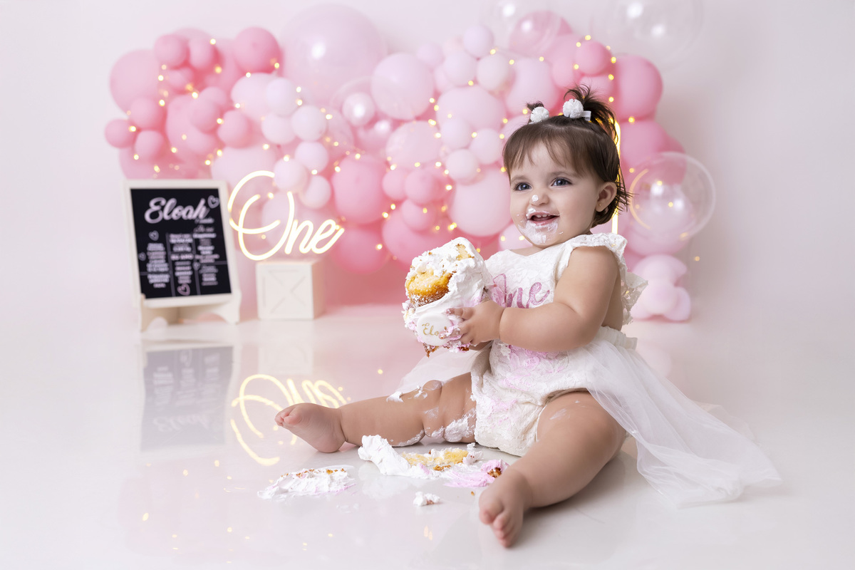 Bebê em ensaio Smash the Cake tema princesa rosa em estúdio fotográfico em Caraguatatuba, cenário delicado, vestido rosa, bolo personalizado e criança sorrindo enquanto brinca com o bolo
