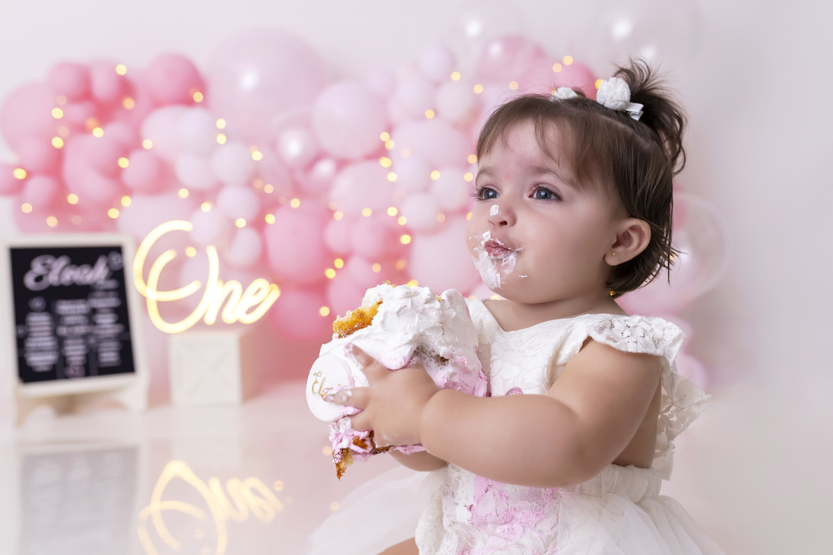 Bebê em ensaio Smash the Cake tema princesa rosa em estúdio fotográfico em Caraguatatuba, cenário delicado, vestido rosa, bolo personalizado e criança sorrindo enquanto brinca com o bolo