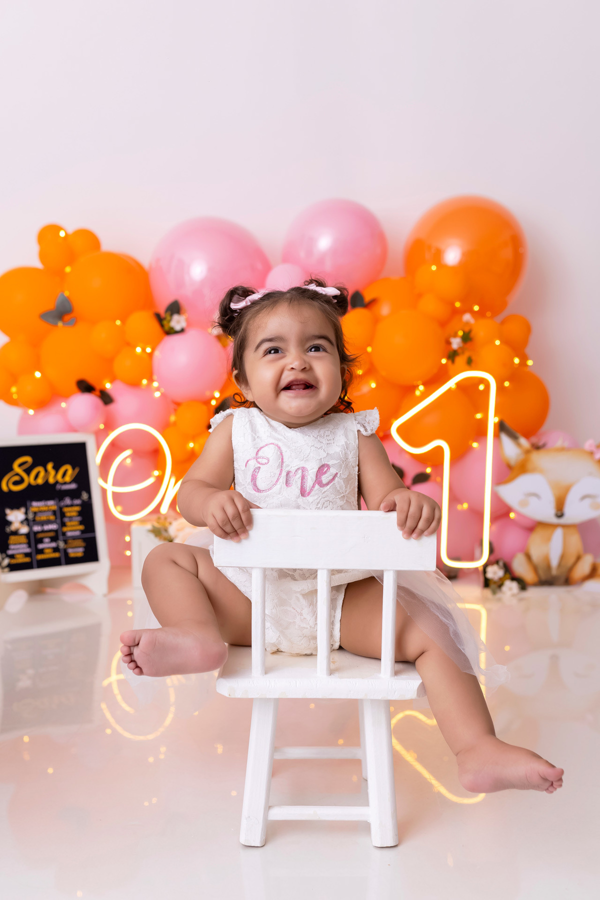 Bebê em ensaio Smash the Cake colorido com flores e raposinha em estúdio em Caraguatatuba, cenário personalizado em tons de rosa e laranja, criança sorrindo e brincando com o bolo de aniversário