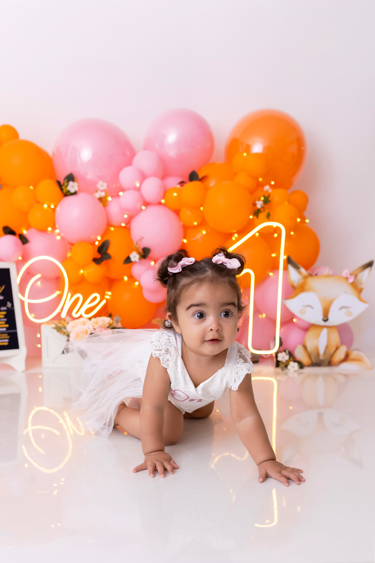 Bebê em ensaio Smash the Cake colorido com flores e raposinha em estúdio em Caraguatatuba, cenário personalizado em tons de rosa e laranja, criança sorrindo e brincando com o bolo de aniversário