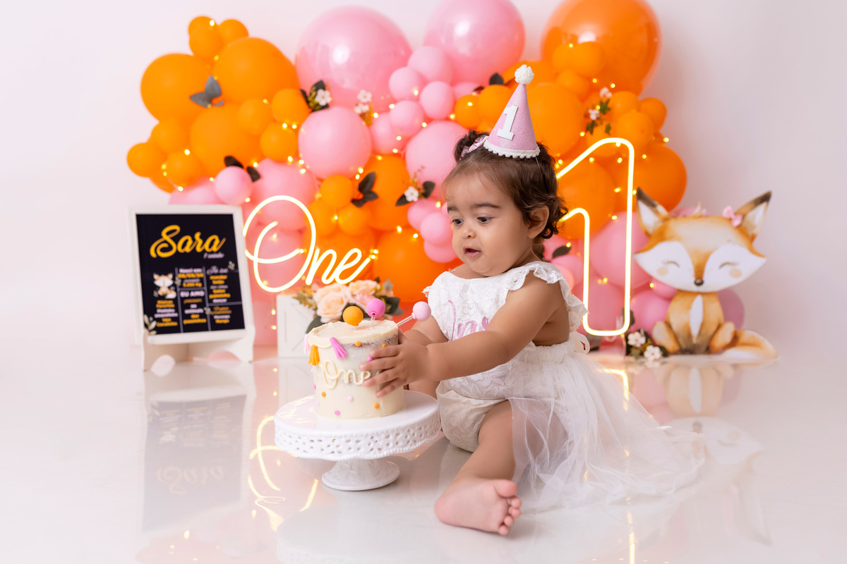 Bebê em ensaio Smash the Cake colorido com flores e raposinha em estúdio em Caraguatatuba, cenário personalizado em tons de rosa e laranja, criança sorrindo e brincando com o bolo de aniversário