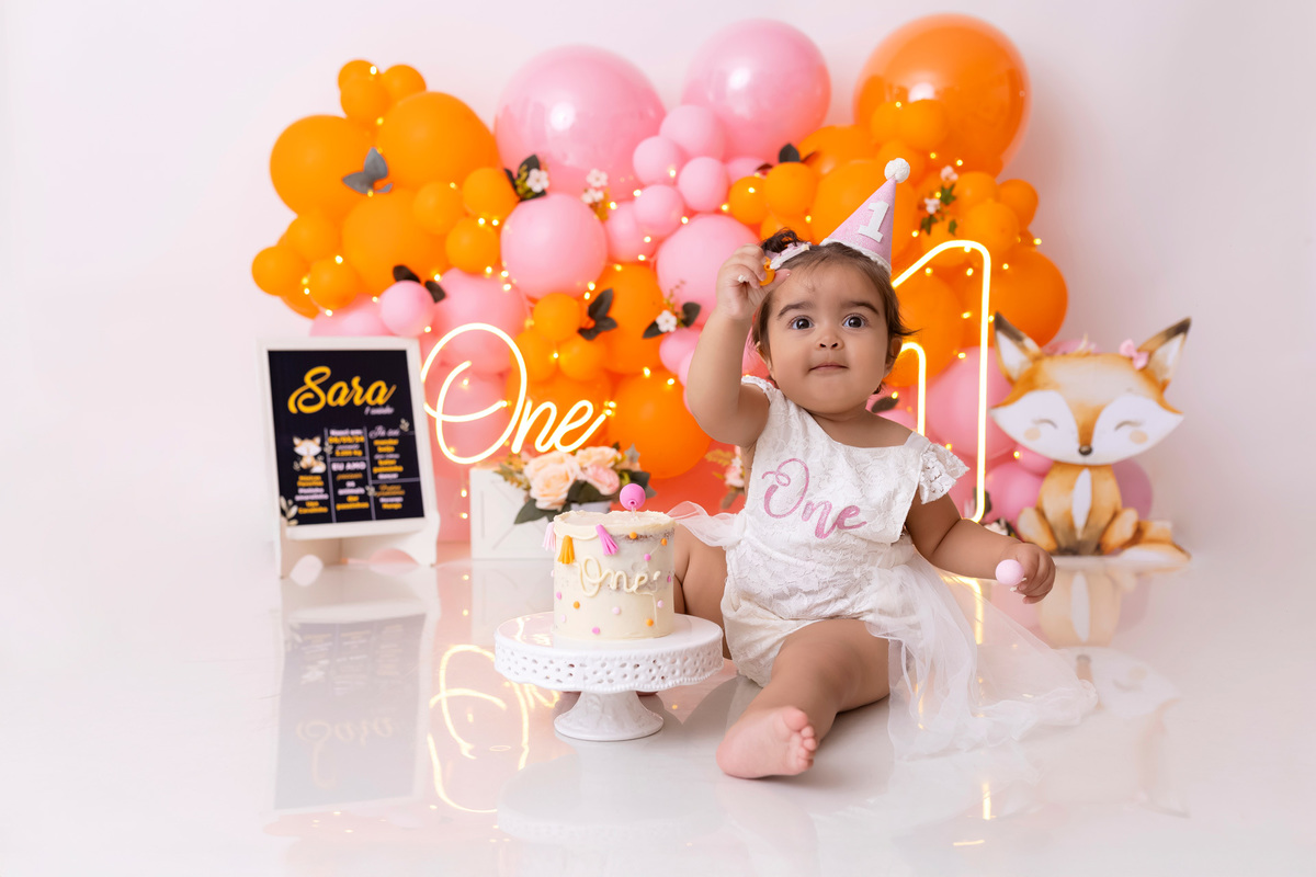Bebê em ensaio Smash the Cake colorido com flores e raposinha em estúdio em Caraguatatuba, cenário personalizado em tons de rosa e laranja, criança sorrindo e brincando com o bolo de aniversário