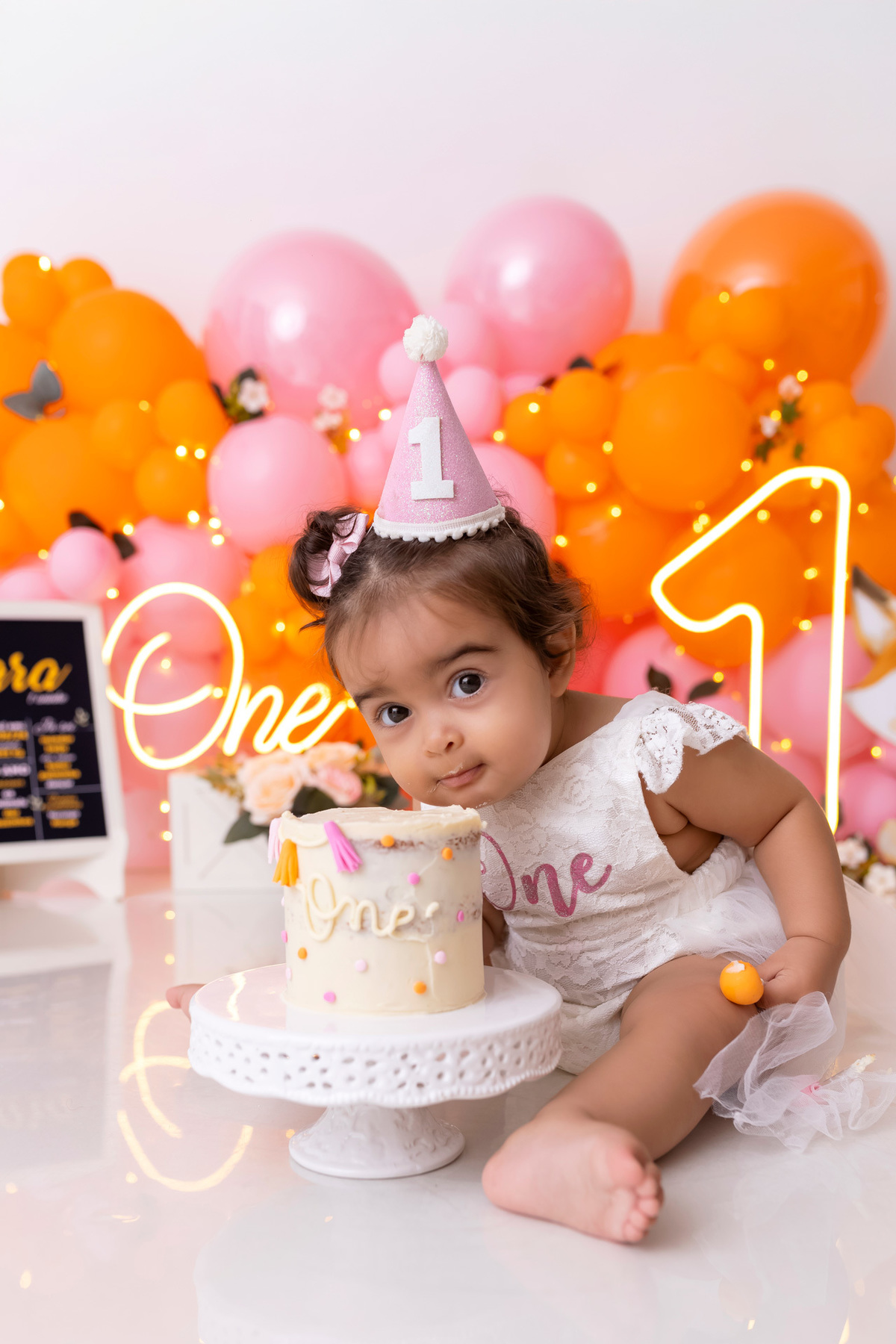 Bebê em ensaio Smash the Cake colorido com flores e raposinha em estúdio em Caraguatatuba, cenário personalizado em tons de rosa e laranja, criança sorrindo e brincando com o bolo de aniversário