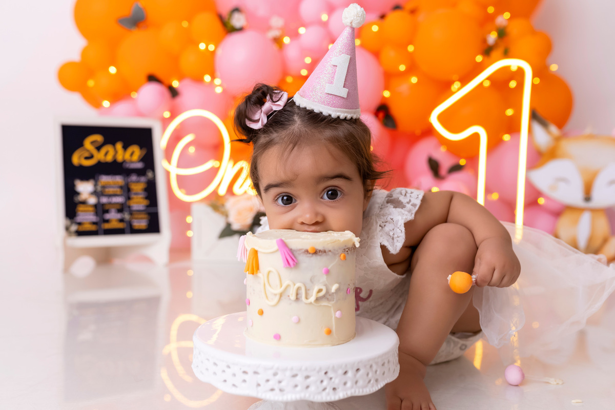 Bebê em ensaio Smash the Cake colorido com flores e raposinha em estúdio em Caraguatatuba, cenário personalizado em tons de rosa e laranja, criança sorrindo e brincando com o bolo de aniversário