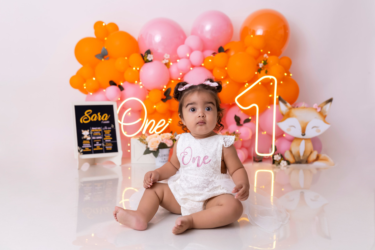 Bebê em ensaio Smash the Cake colorido com flores e raposinha em estúdio em Caraguatatuba, cenário personalizado em tons de rosa e laranja, criança sorrindo e brincando com o bolo de aniversário