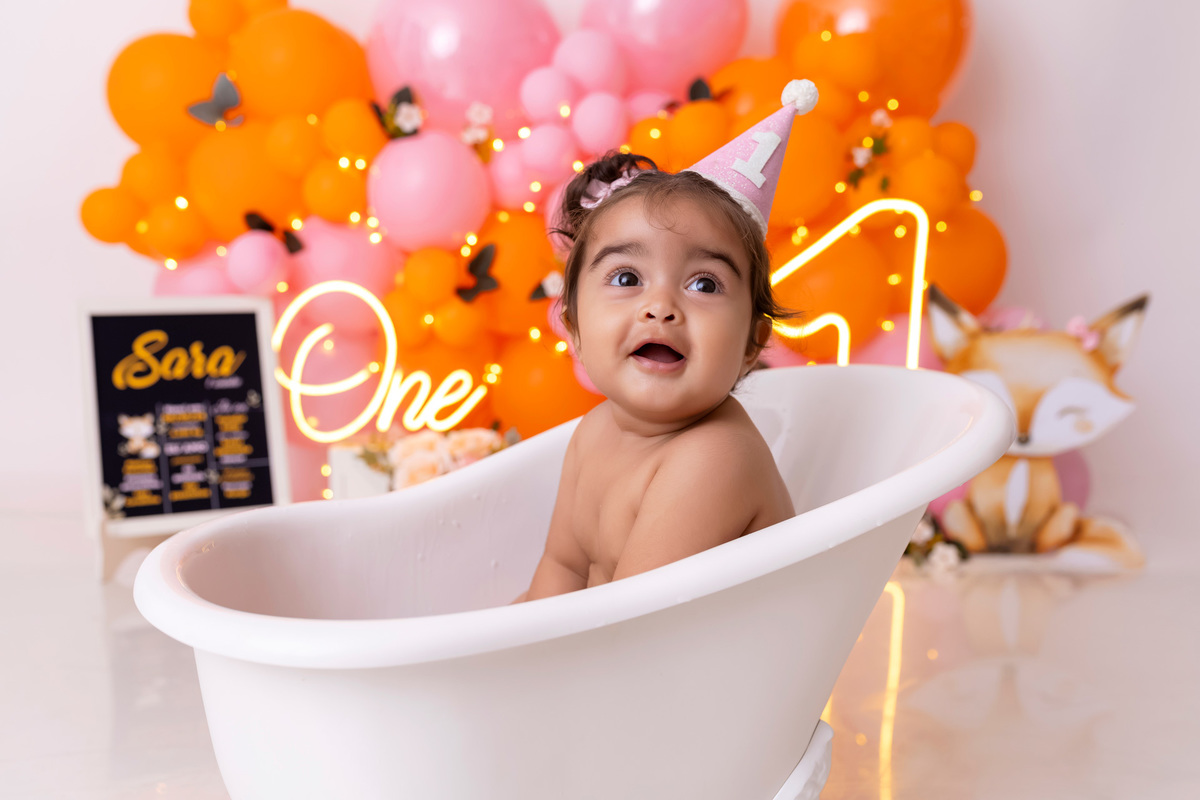 Bebê em ensaio Smash the Cake colorido com flores e raposinha em estúdio em Caraguatatuba, cenário personalizado em tons de rosa e laranja, criança sorrindo e brincando com o bolo de aniversário