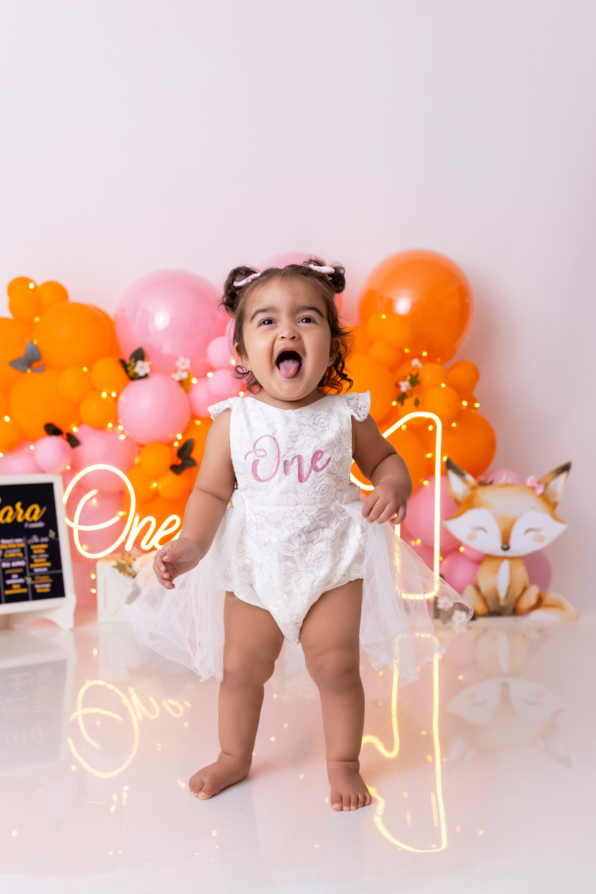 Bebê em ensaio Smash the Cake colorido com flores e raposinha em estúdio em Caraguatatuba, cenário personalizado em tons de rosa e laranja, criança sorrindo e brincando com o bolo de aniversário