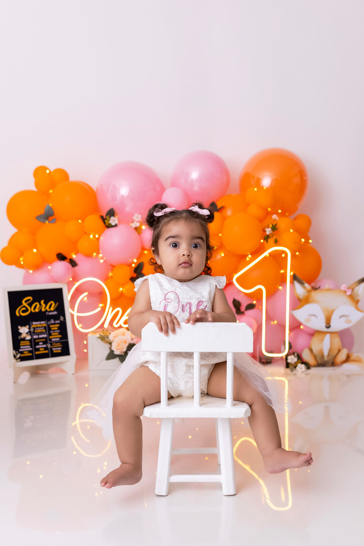 Bebê em ensaio Smash the Cake colorido com flores e raposinha em estúdio em Caraguatatuba, cenário personalizado em tons de rosa e laranja, criança sorrindo e brincando com o bolo de aniversário