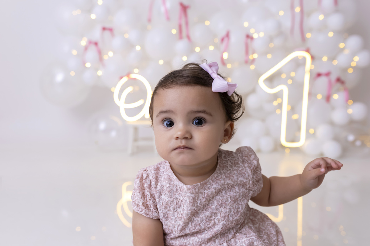 Bebê em ensaio Smash the Cake clean em estúdio branco com laços rosa, cenário minimalista e elegante, criança comendo e se lambuzando com bolo de aniversário em Caraguatatuba