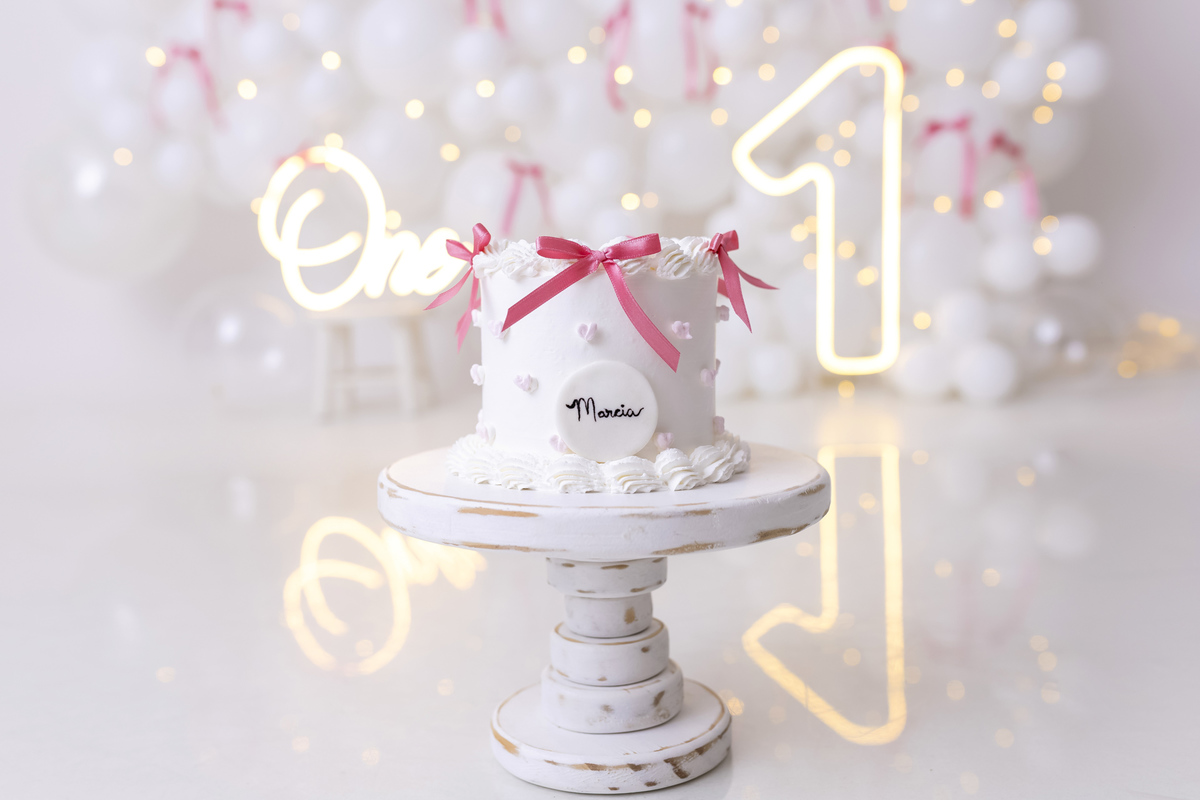 Bebê em ensaio Smash the Cake clean em estúdio branco com laços rosa, cenário minimalista e elegante, criança comendo e se lambuzando com bolo de aniversário em Caraguatatuba