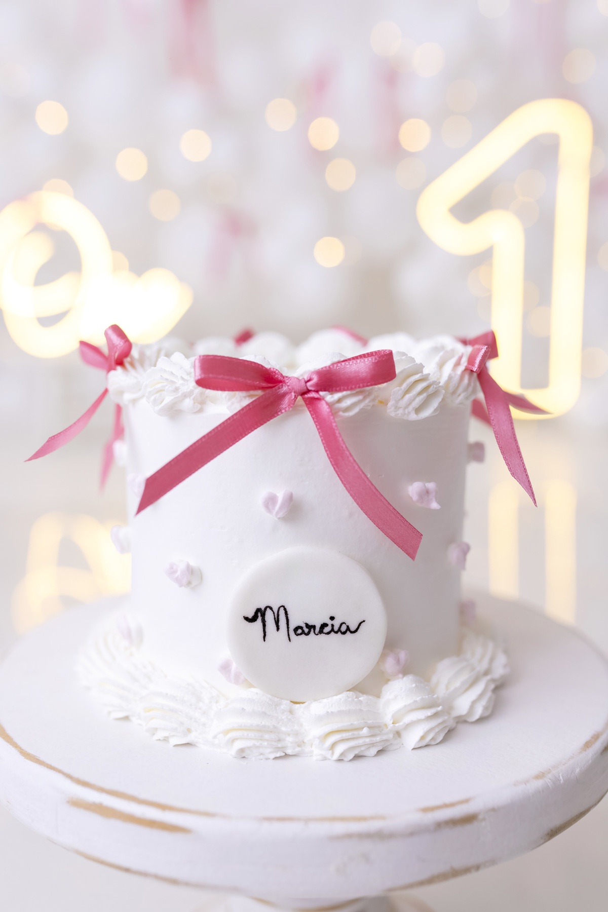 Bebê em ensaio Smash the Cake clean em estúdio branco com laços rosa, cenário minimalista e elegante, criança comendo e se lambuzando com bolo de aniversário em Caraguatatuba