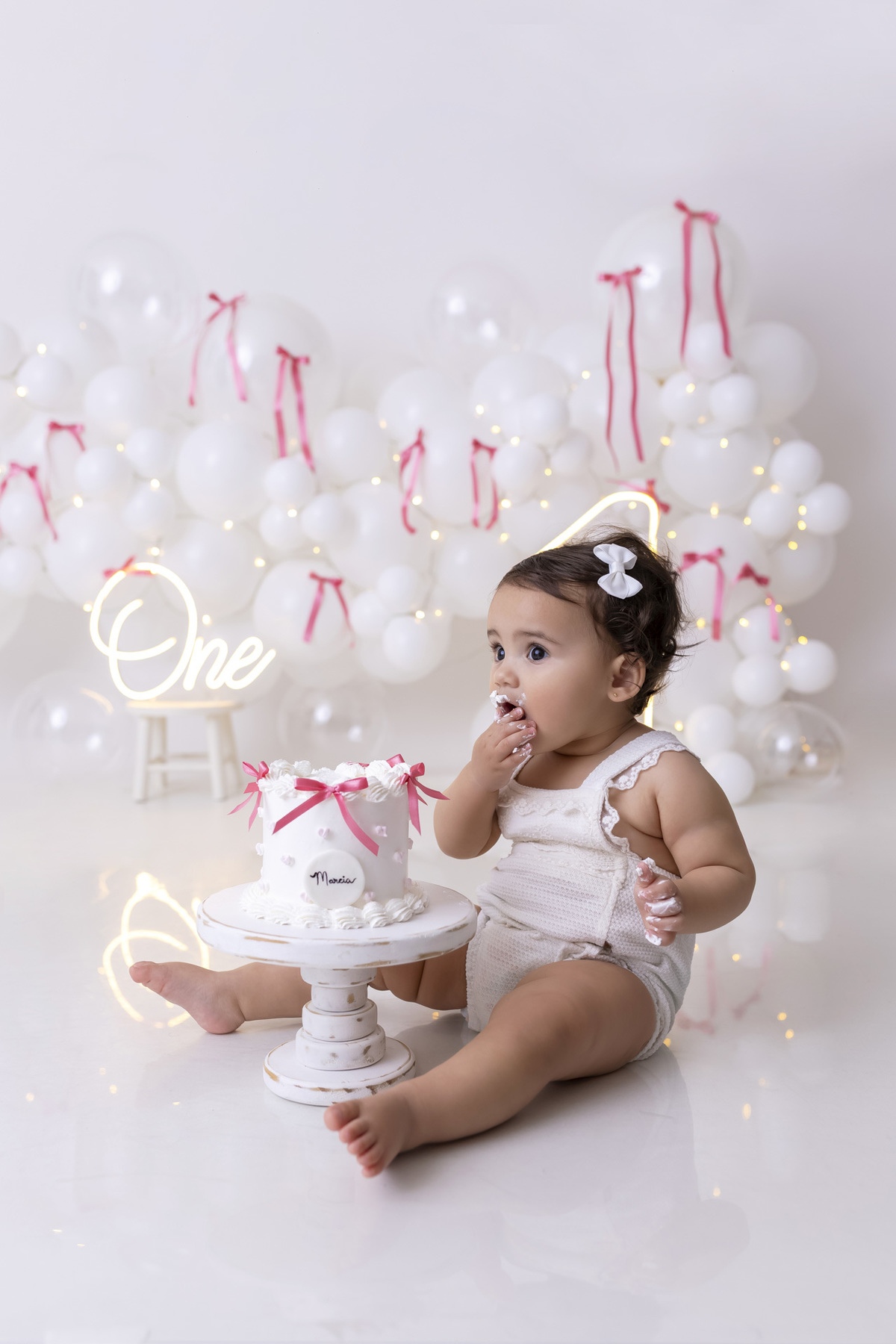 Bebê em ensaio Smash the Cake clean em estúdio branco com laços rosa, cenário minimalista e elegante, criança comendo e se lambuzando com bolo de aniversário em Caraguatatuba