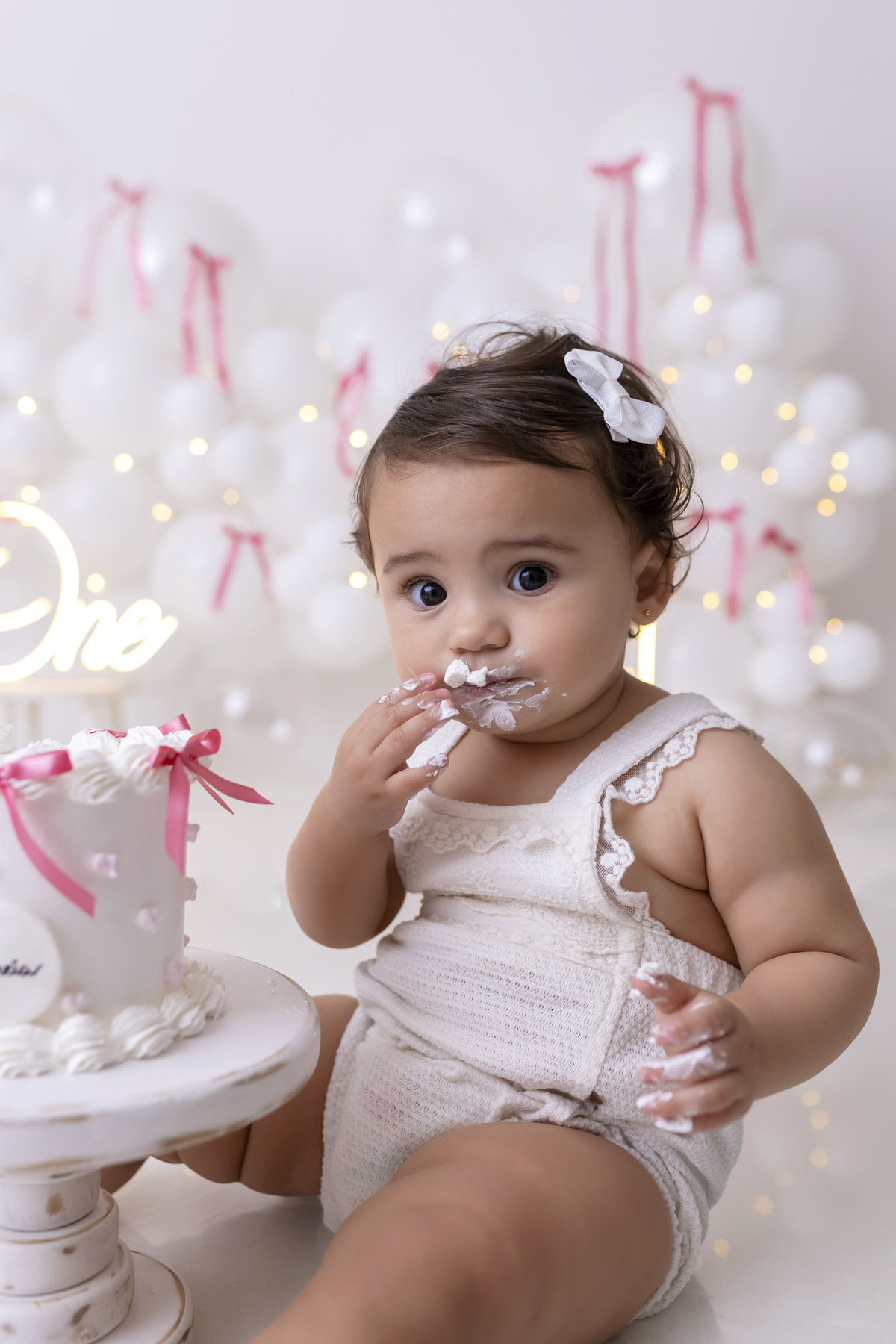 Bebê em ensaio Smash the Cake clean em estúdio branco com laços rosa, cenário minimalista e elegante, criança comendo e se lambuzando com bolo de aniversário em Caraguatatuba