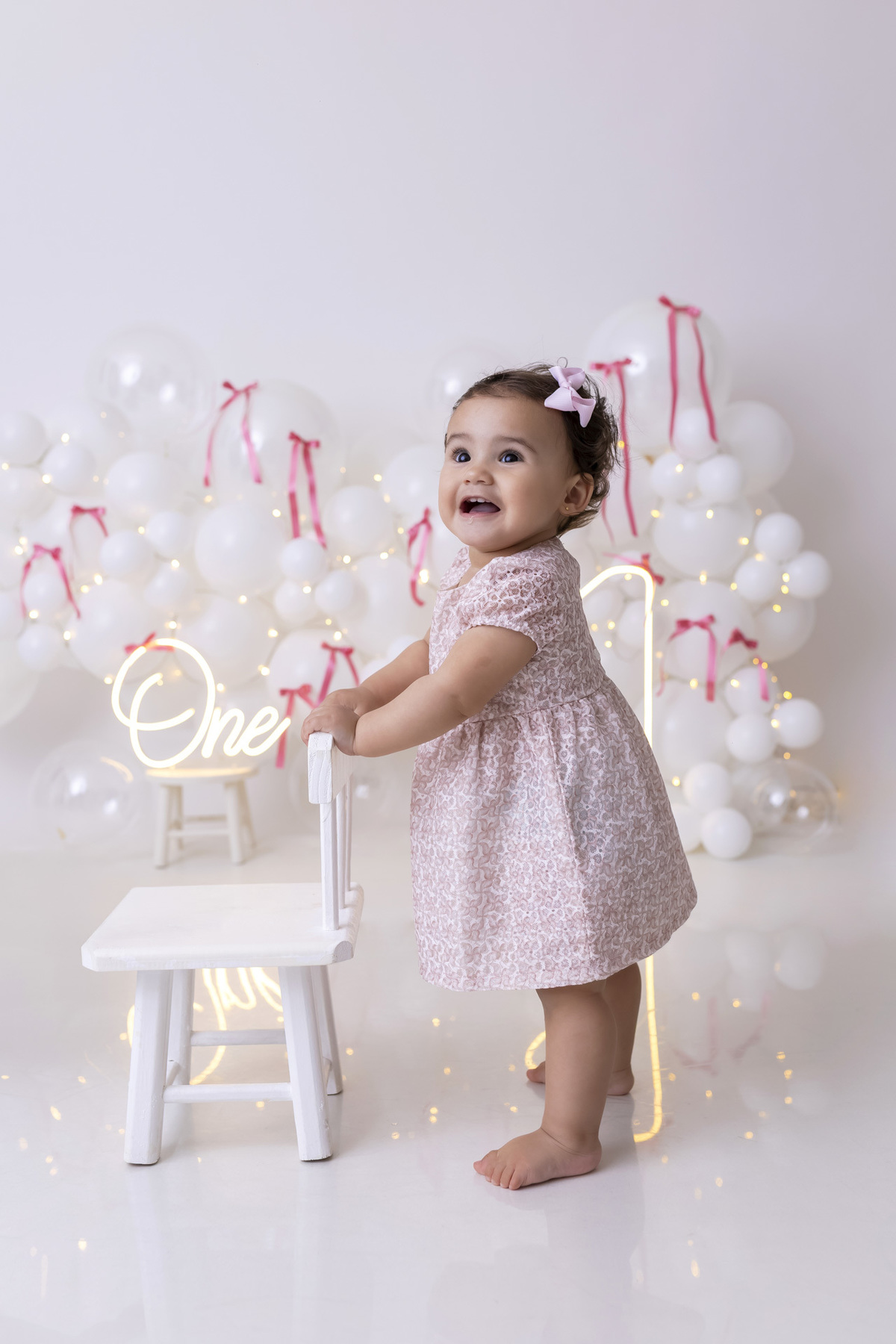 Bebê em ensaio Smash the Cake clean em estúdio branco com laços rosa, cenário minimalista e elegante, criança comendo e se lambuzando com bolo de aniversário em Caraguatatuba