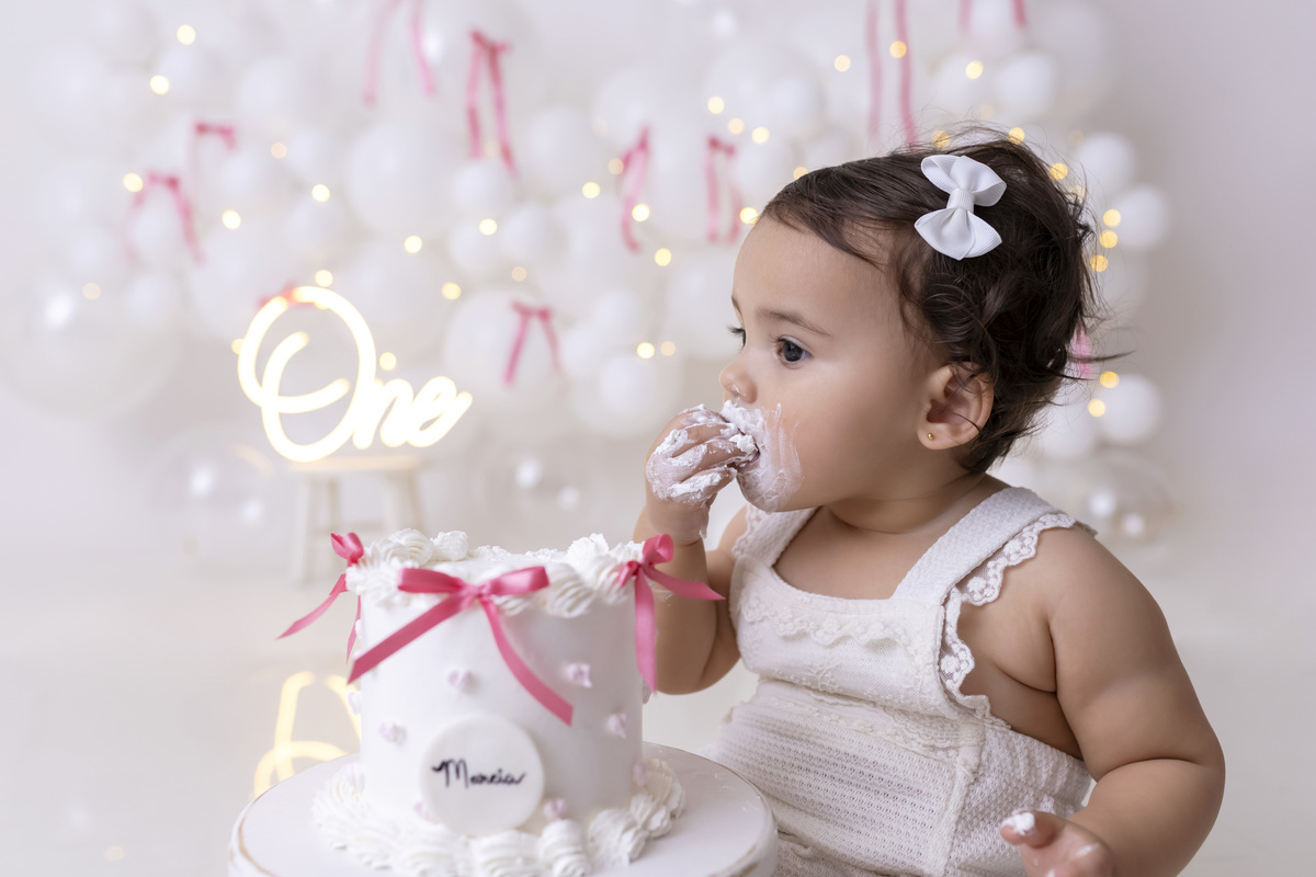 Bebê em ensaio Smash the Cake clean em estúdio branco com laços rosa, cenário minimalista e elegante, criança comendo e se lambuzando com bolo de aniversário em Caraguatatuba