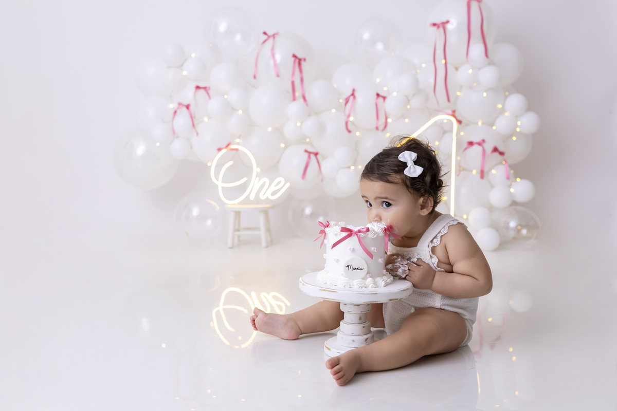 Bebê em ensaio Smash the Cake clean em estúdio branco com laços rosa, cenário minimalista e elegante, criança comendo e se lambuzando com bolo de aniversário em Caraguatatuba