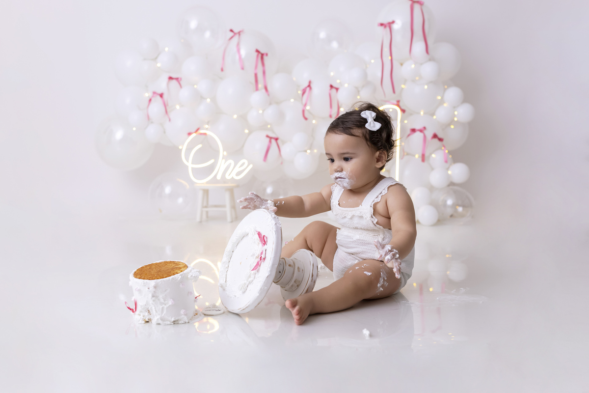 Bebê em ensaio Smash the Cake clean em estúdio branco com laços rosa, cenário minimalista e elegante, criança comendo e se lambuzando com bolo de aniversário em Caraguatatuba